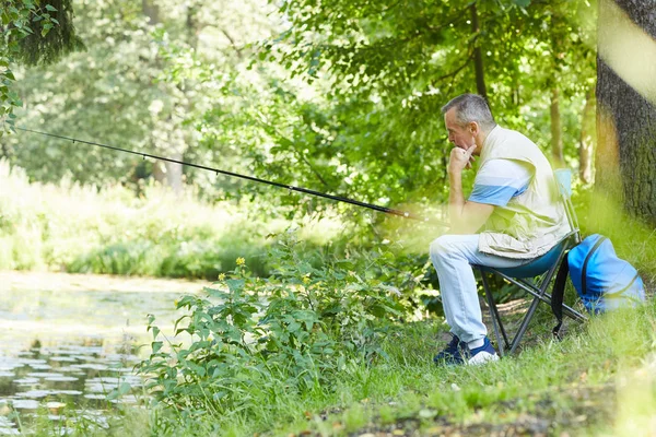 Mature fisherman sitting on chair with fishing rod fishing at the lakeside in nature