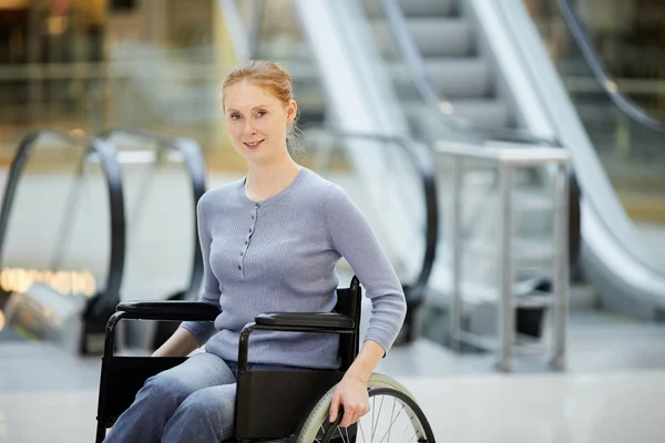 Portrait of young disabled woman in wheelchair smiling at camera while visiting shopping mall with escalator in background