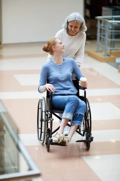 坐在轮椅上与年迈的母亲一起在购物中心散步的残疾青年妇女 — 图库照片