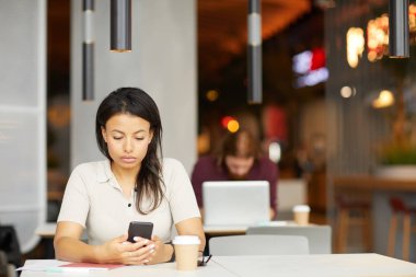 Ciddi genç bir kadın masada oturuyor ve kafede arka planda bir adamla internet üzerinden çalışmak için cep telefonu kullanıyor.