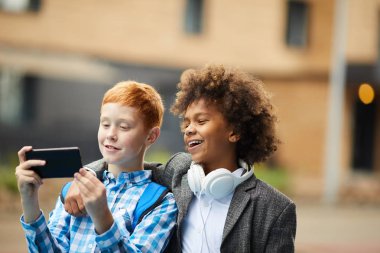 Dışarıda cep telefonunda selfie çekerken kameraya sarılıp gülümseyen iki çocuk.