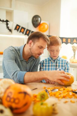 Genç baba, evdeki masada otururken oğluna balkabaklarından süslemeler yaptığını anlatıyor.