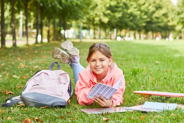 かわいい女の子の肖像画緑の草の上に横たわるバックパックと本と読書前に学校でのレッスン — ストック写真