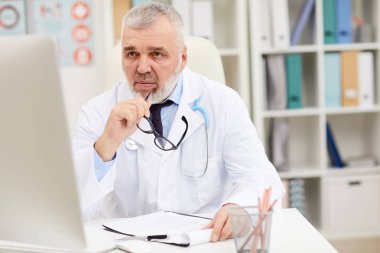 Gri saçlı ve sakallı olgun bir doktor masada oturuyor ve ofiste çalışırken belge okuyor.