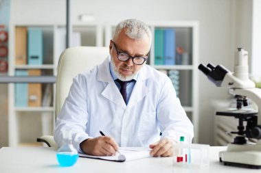 Laboratuvardaki tıbbi araştırmalar sırasında beyaz önlüklü olgun bir doktorun masaya oturup panoya yazı yazması. 