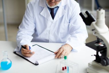 Laboratuvardaki tıbbi araştırmalar sırasında beyaz önlüklü olgun bir doktorun masaya oturup panoya yazı yazması. 