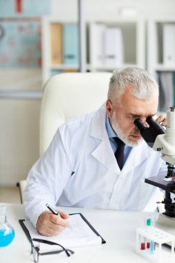 Ofisteki bilimsel araştırmalar sırasında mikroskoba bakan gri saçlı kıdemli doktorun yakın çekimi.
