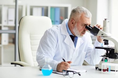 Masada oturup ofisteki mikroskoptan bakmaya odaklanmış ciddi bir yetişkin bilim adamı. 
