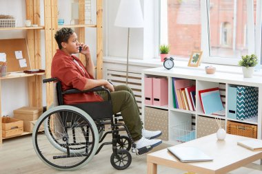 Engelli Afrikalı genç bir kadın tekerlekli sandalyede oturmuş evdeki cep telefonuyla konuşuyor.