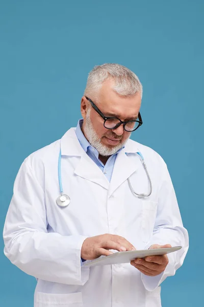 白髪と白のコートを持つシニア医師の肖像画は 青の背景にタブレットPc上でオンラインで動作 — ストック写真