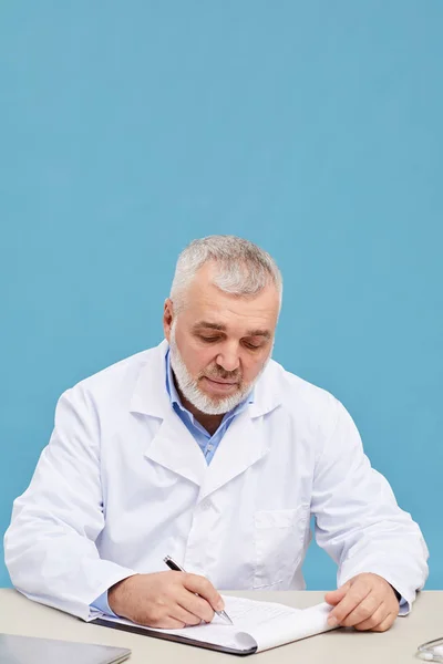青の背景に医療カードを埋める職場で働く白髪とひげを持つ熟女医師 — ストック写真