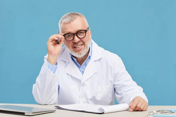 灰色の髪とひげを持つ成熟した医師の肖像画とテーブルで文書を扱う眼鏡 — ストック写真