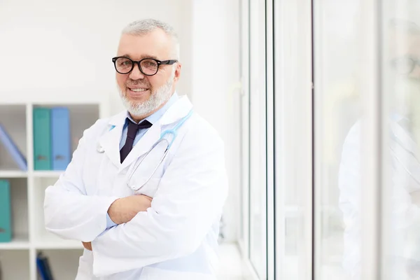 眼鏡のシニア医療専門家の肖像画や腕を組んで立つ白衣姿が交差し 病院のカメラで笑顔 — ストック写真