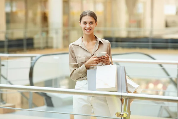 Smiling young woman holding paper bags and using mobile phone while walking in the shopping mall