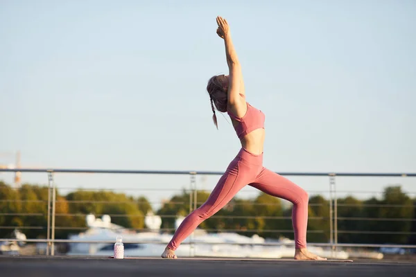 Young woman in pink sports clothing standing and stretching hands up practicing yoga in the city