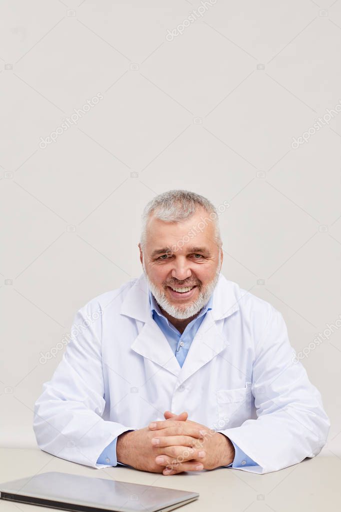 Portrait of mature doctor with grey hair and in white coat smiling at camera while sitting at workplace over white background