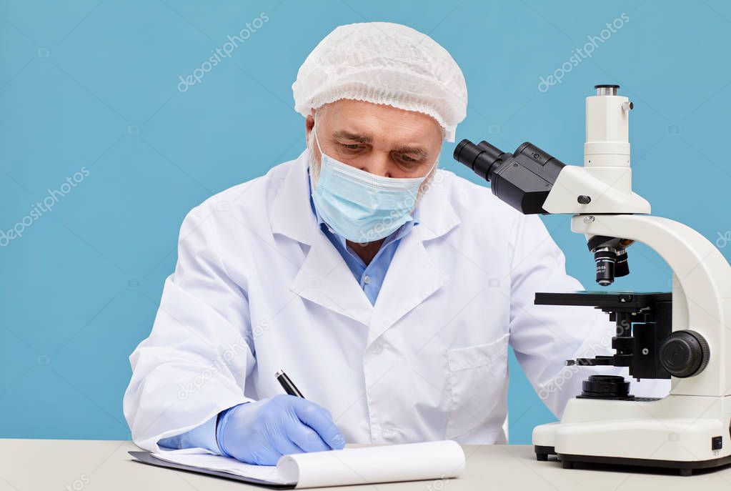 Serious chemist in protective workwear sitting at the table with microscope and making notes in document on blue background