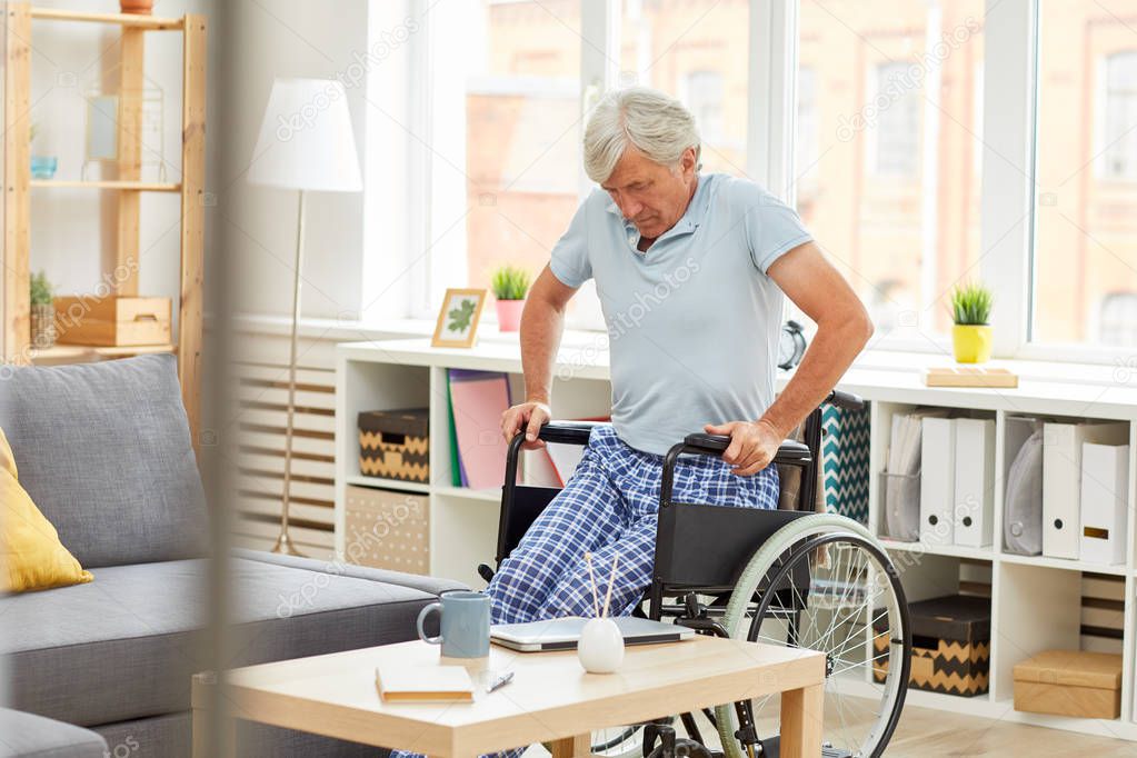 Senior man with grey hair sitting in wheelchair while resting in the room at home
