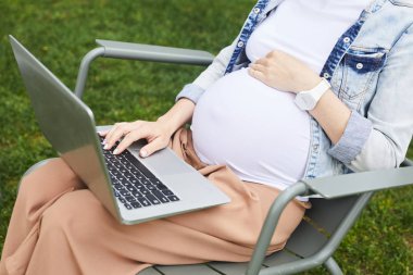 Genç hamile bir kadının sandalyede oturup dizüstü bilgisayarda temiz havada yazışının yakın çekimi.