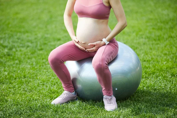 Young pregnant woman in pink sport clothing sitting on fitness ball and doing exercises on fresh air