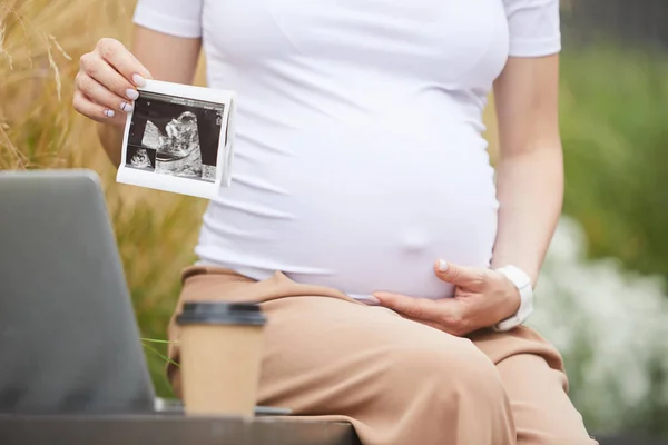 孕妇坐在长椅上 拿着笔记本电脑和咖啡饮料 抱着婴儿超声波的特写镜头 — 图库照片