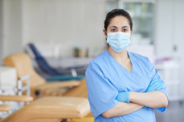 Maskeli ve üniformalı hemşirenin portresi kollarını kavuşturup hastaneye bakıyor.