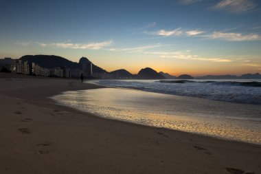 Sabahın erken saatlerinde Rio de Janeiro'daki Copacabana plajı, gün doğumundan hemen önce arka planda Sugarloaf dağı ile gökyüzünde derin turuncu ve mavi renklerle