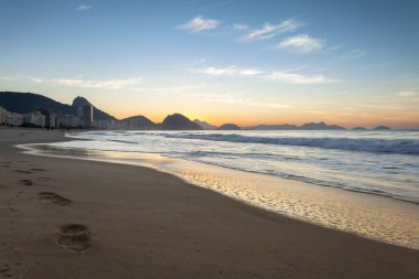 Rio de Janeiro Copacabana plajda okyanus dalgaları boyunca Ayak Sesleri sabahın erken saatlerinde arka planda Sugarloaf dağ ile turuncu ve mavi gündoğumu