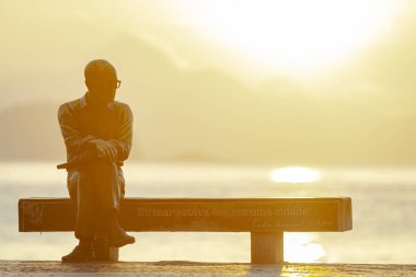 Brezilyalı şair Carlos Drummond de Andrade Rio de Janeiro heykeli Copacabana bulvarı üzerinde bir bankta oturan okyanus ve Itroi arka bir gündoğumu ile arkadan arkadan arkadan bir gündoğumu ile arkadan aydınlatılmış
