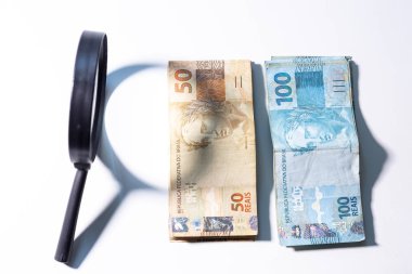 Yanında bir büyüteç ile Brezilya Real banknotlar Yığını. Temiz stüdyo beyaz bir arka plan karşı büyütme aracı ile para vurdu.