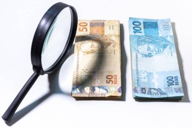 Yanında bir büyüteç ile Brezilya Real banknotlar Yığını. Temiz stüdyo beyaz bir arka plan karşı büyütme aracı ile para vurdu.