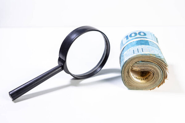 Thick roll of Brazilian Real banknotes next to a magnifying glass with soft shadow. Clean studio shot currency with magnify tool against a white background.