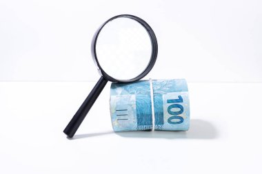 Brezilya Reali banknotkalın rulo bir büyüteç altında yüz değeri ileriye bakan yan yana aydınlatılmış. Temiz stüdyo beyaz bir arka plan karşı büyütme aracı ile para vurdu.