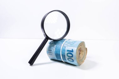 Brezilya Reali banknotkalın rulo bir büyüteç altında yüz değeri ileriye bakan yan yana aydınlatılmış. Temiz stüdyo beyaz bir arka plan karşı büyütme aracı ile para vurdu.