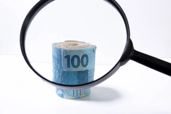 Focus on the top of a thick roll of banknotes seen through a magnifying glass. Clean studio shot of currency with magnify tool against a white background.