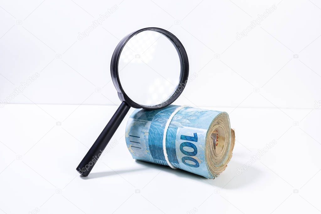 Thick roll of Brazilian Real banknotes below a magnifying glass lit from the side with the one hundred value facing forward. Clean studio shot currency with magnify tool against a white background.