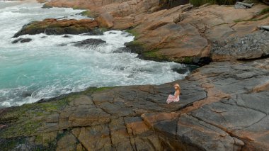 Okyanusa bakan büyük bir kıyı kayasında meditasyon pozisyonunda şapkalı kadın gün doğumunda yavaşça içeri dalıyor.