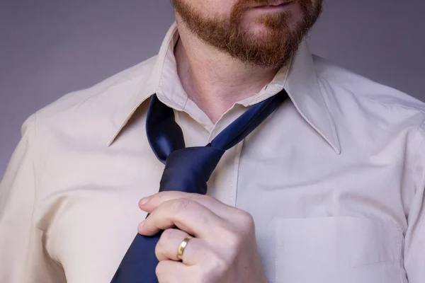 Close up of red bearded married man tying his blue tie over bright shirt twisting his neck looking off to the right on grey studio background