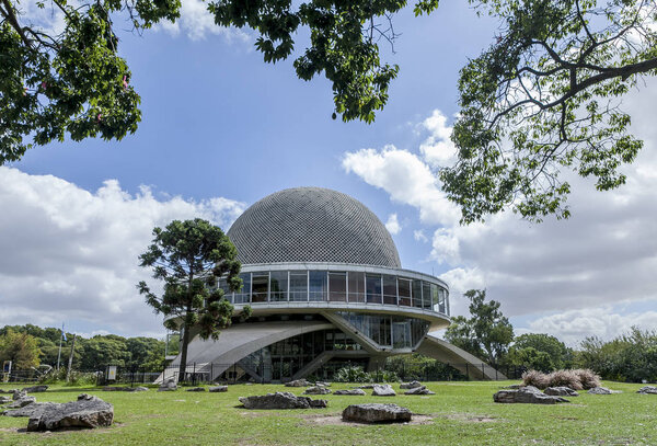 Buenos Aires, Argentina - March 30, 2012: The planetarium of Buenos Aires, Argentina framed by tree branches with rocks in the field in front of it and clouds in the blue sky