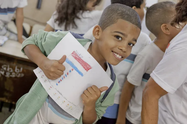 巴西里约热内卢 2015年7月2日 巴西小男孩拿着一张画 面带微笑 对着镜头竖起两个大拇指 背景是同龄人 — 图库照片