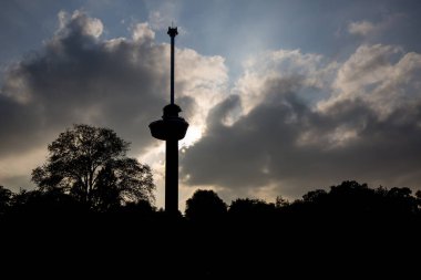 Rotterdam 'da siluetteki bir Euromast 'ın yatay görüntüsü, Hollanda, muhteşem bir sahne oluşturan bulutların arkasındaki güneşle gün batımında Şehir Parkı üzerinde yükselen.