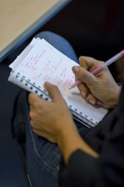 Left hand holding a notebook on a female leg with blue jeans on while writing notes with the right hand with a pink ink pen. Vertical shot of someone writing on lap.