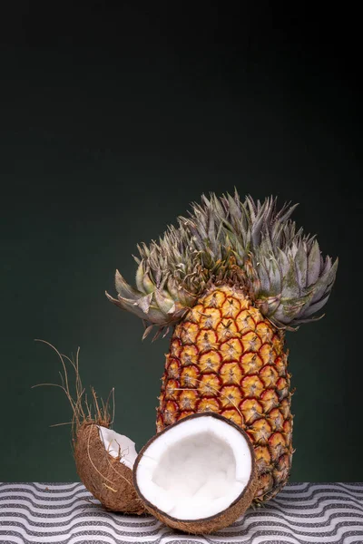 ココナッツの2つの半分の後ろに面白いトップを持つ特徴的なパイナップルは 暗い緑色の背景に対してパターン化された表面に外側の茶色の殻と内側の白いココを示しています — ストック写真