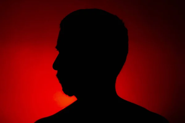 Cleanly defined silhouette of a male person turned to the left against a red background with a spotlight and bright area right behind the bust. Studio shot with well defined colour background.