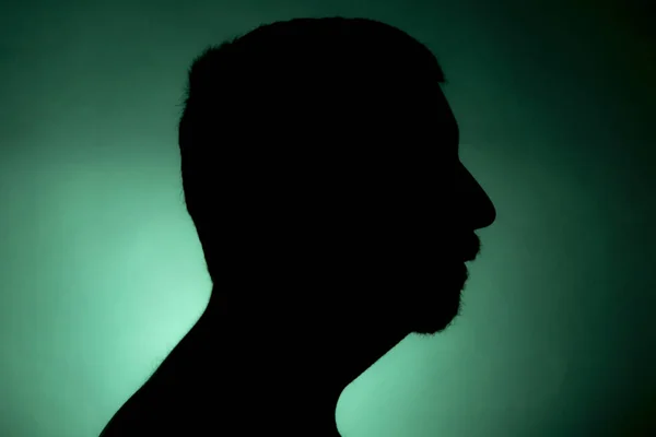 Cleanly defined silhouette of a male person turned to the right against a green background with a spotlight and bright area right behind the bust. Studio shot with strong explicit colour background.