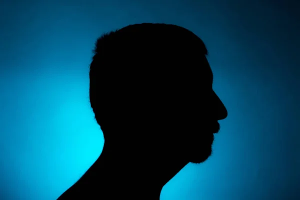 Cleanly defined silhouette of a male person turned to the right against a blue background with a spotlight and bright area right behind the bust. Studio shot with strong explicit colour background.