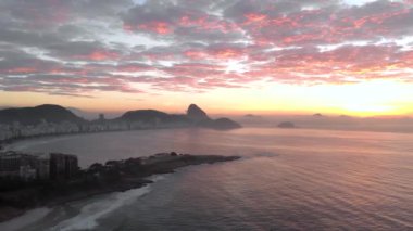 Rio de Janeiro'daki Arpoador kayasını gösteren hava rotasyon tavası, altın bir gündoğumu yla renkli bir şekilde aydınlatarak Copacabana'nın göbeğini ve mahallesini ve Sugarloaf dağının arka planda olduğunu ortaya çıkardı