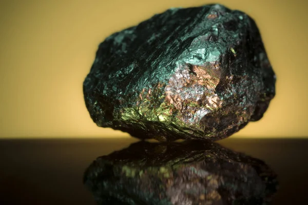 Small piece of heavy mineral rock under a green light source that has gold features shining through against an orange background