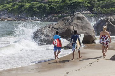 Rio de Janeiro, Brezilya - 30 Ocak 2016: Sörf tahtalı iki erkek ve sahilde yürüyen bir kadın arka planda kayalara çarpan dalgalar geçiyor