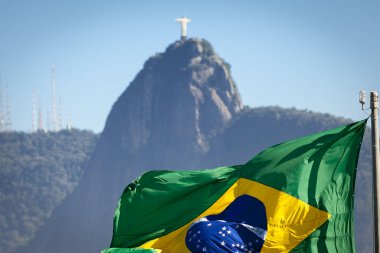 Mavi bir gökyüzü ile güneşli bir günde ön planda odak renkli bir Brezilya bayrağı ile arka planda ünlü Mesih heykeli birleştiren